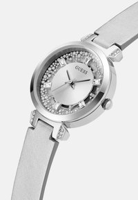 Sølvtonet ur med en rund kasse med krystal detaljer, et struktureret hvidt læderrem og en glitrende bezel omkring en sølvskive.