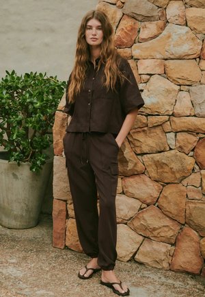 Junge Frau mit langen welligen Haaren steht vor einer Steinmauer, trägt ein lockeres dunkles Hemd mit Knöpfen, passende Hose und schwarze Flip-Flops.