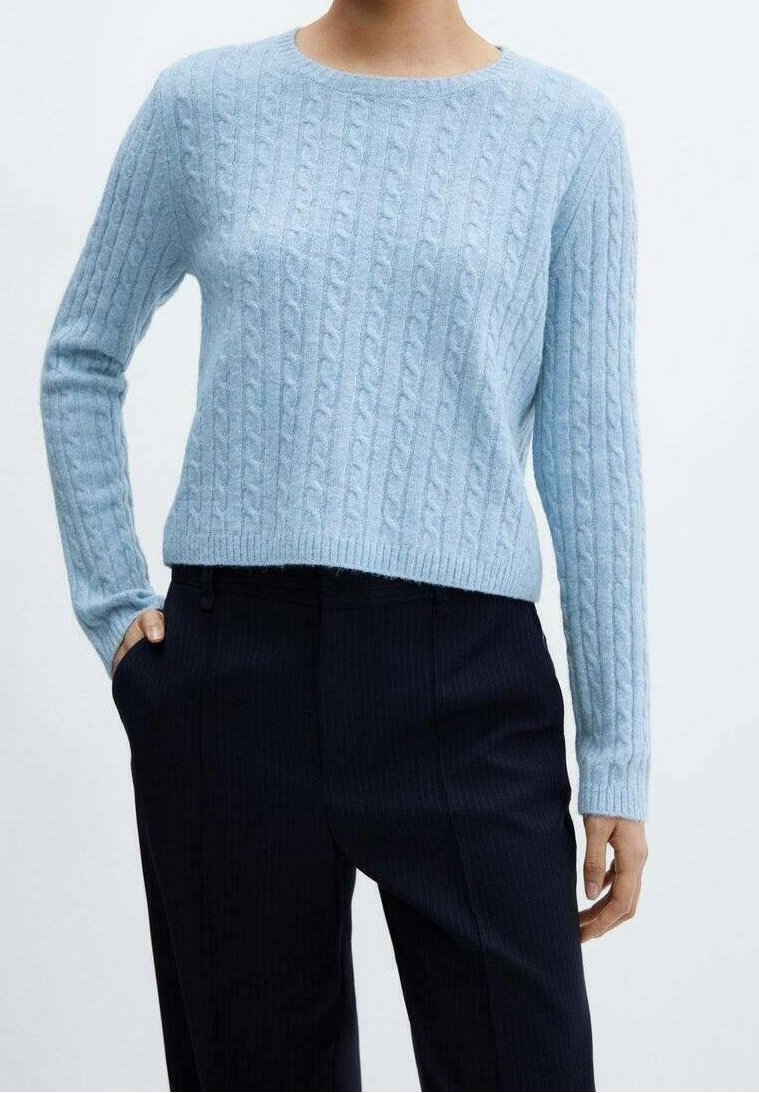 Personne portant un pull torsadé bleu clair et un pantalon noir, une main dans la poche, sur un fond blanc uni.