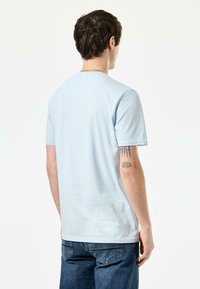 Lichtblauw T-shirt met korte mouwen van zachte katoen, met een ronde hals en een losse pasvorm. Zichtbare tatoeage op de onderarm, gedragen met een spijkerbroek.