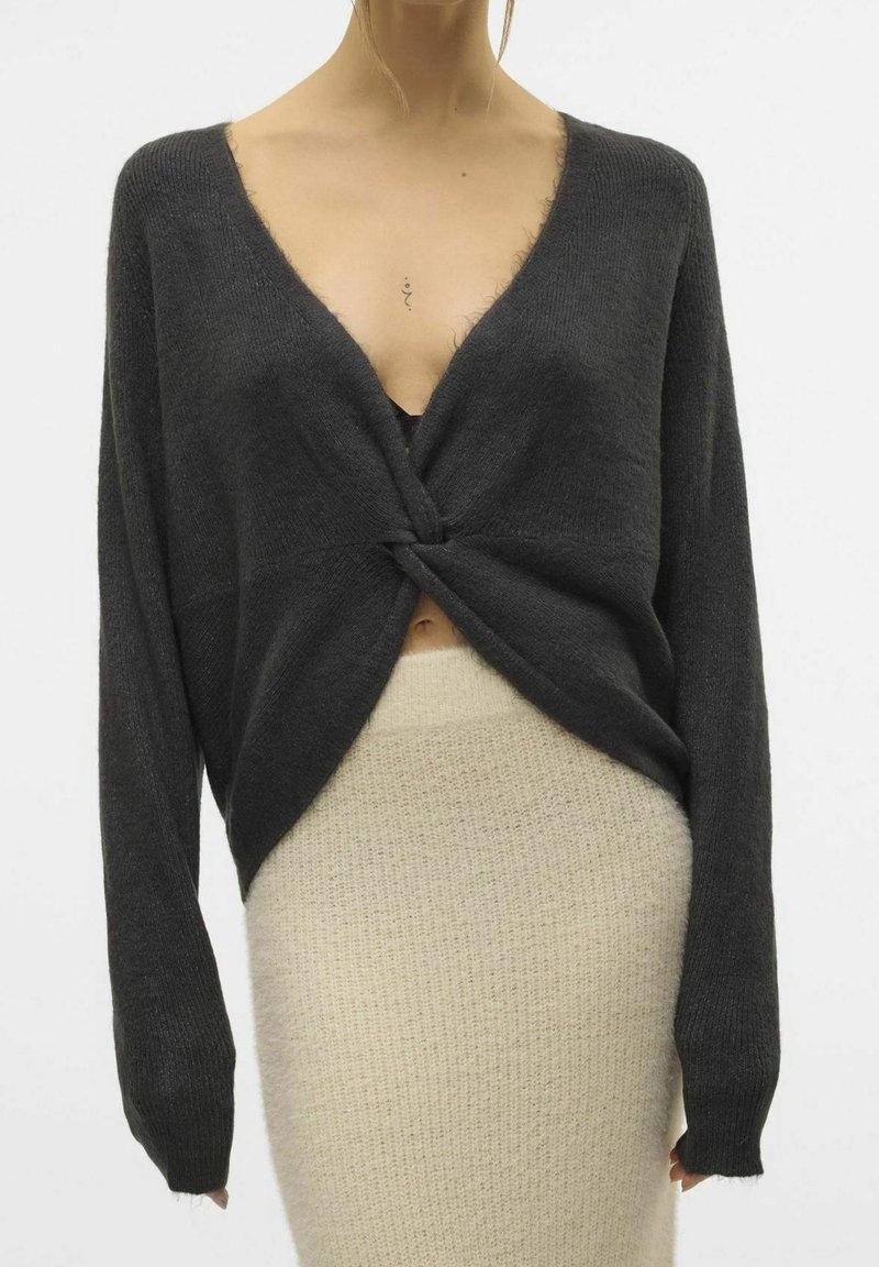 Femme portant un pull à col en V torsadé gris foncé et une jupe en maille crème taille haute sur un fond uni.