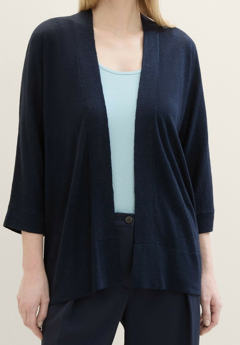 Femme portant un cardigan bleu marine ample et ouvert sur le devant, par-dessus un débardeur bleu clair, associé à un pantalon bleu marine, sur un fond uni.