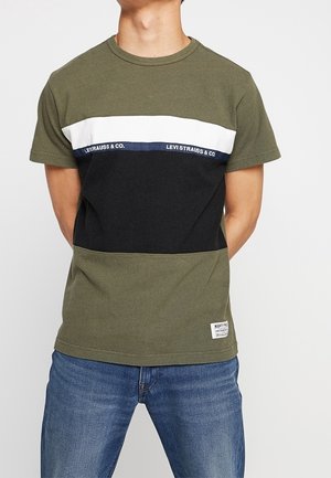 Short-sleeve t-shirt in olive green, featuring a horizontal black and white stripe across the chest with branding. Soft cotton material.