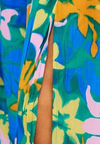 Φωτεινό υφασμάτινο με ανθικά μοτίβα σε κίτρινο, πράσινο, μπλε και ροζ χρώμα· διαθέτει κάθετη σχισμή που αποκαλύπτει μαυρισμένο δέρμα από κάτω.