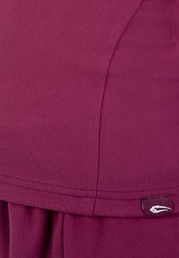 Nahaufnahme eines kastanienbraunen Stoffs mit sichtbarer Naht und kleinem schwarzem Etikett mit weißem Logo unten rechts.