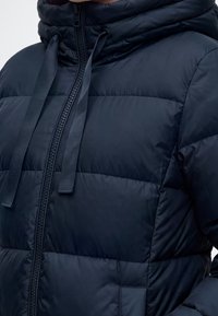 Κοντινό πλάνο ενός ατόμου που φοράει μπλε σκούρα καπιτονέ μπουφάν με φερμουάρ μπροστά και κουκούλα με κορδόνι.