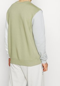 Man i en grön och grå färgblockerad sweatshirt och ljusa byxor, som vänder ryggen mot kameran mot en enfärgad bakgrund.