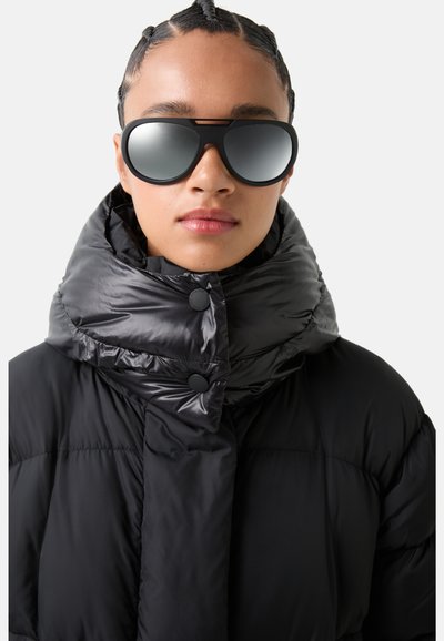 Black puffer jacket with a high collar, featuring glossy fabric and two black buttons. Large black sunglasses with mirrored lenses.