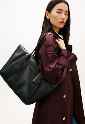 Femme portant un manteau bordeaux tenant un grand sac fourre-tout noir avec une ferrure dorée sur l'épaule, sur fond blanc.
