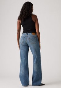 Jeans de mezclilla azules con cintura alta y corte de pierna ancha. Presenta bolsillos traseros y un parche de cuero con el logo de la marca. Textura de lavado desgastado.
