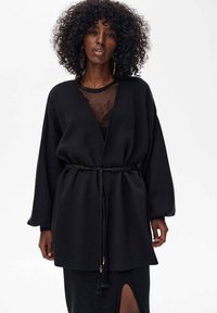 Czarny, luźny płaszcz o szerokich rękawach, z detalem w postaci paska, miękka faktura, noszony na przejrzystej bluzce. Płaszcz ozdobiony frędzlami na końcach paska.