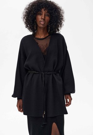 Czarny, luźny płaszcz o szerokich rękawach, z detalem w postaci paska, miękka faktura, noszony na przejrzystej bluzce. Płaszcz ozdobiony frędzlami na końcach paska.
