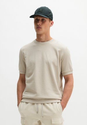 Man draagt een licht beige kortermig overhemd en bijpassende broek met zakken, zwarte pet met logo, staat met handen in de zakken.