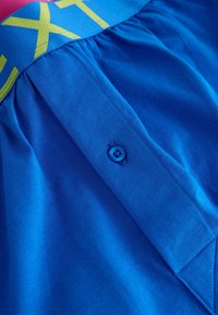 Vêtement en coton bleu avec une ceinture ornée de grands textes jaunes, un détail de bouton, et un tissu froncé pour une apparence fluide.