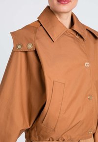 Giacca corta marrone con ampio colletto, due dettagli in metallo dorato sulle spalle e una tasca frontale. Tessuto dalla trama liscia.