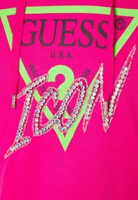 Sudadera de color rosa brillante con un logo triangular verde, "GUESS," y la palabra "TOON" adornada con brillantes de strass. Tejido texturizado.