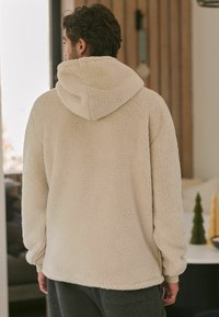 Beige fleece-huppari, jossa on rakenne ja pörröinen pinta, varustettuna suurella hupulla ja rennolla istuvuudella. Hihansuut ja helma on viimeistelty sileällä reunalla.