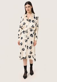 Rochie midi florală de culoare crem cu modele de flori negre, cu nasturi pe față, mâneci lungi și un brâu legat. Pur tată cu ciorapi negri transparenți și pantofi ascuțiți.