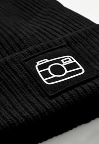 Black knit beanie featuring a textured pattern and a rectangular black patch with a white camera graphic logo.