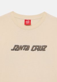 Iekšējo īsām piedurknēm beidž t-krekls ar apaļu apkakli, uz kura priekšpuses ir brūns, blokveida "JANTA CRUZ" logotips. Kokvilnas materiāls, mīksta tekstūra.