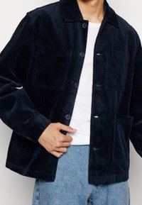 Chaqueta de pana azul marino con cuello tipo solapa, cierre frontal de botones, dos bolsillos en el pecho y textura acanalada. Llevada sobre una camisa blanca.