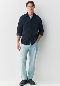 Mørkeblå skjorte med knapper og to brystlommer, lyseblå jeans og sorte sko. Glat stof, klassisk krave og oprullede ærmer.