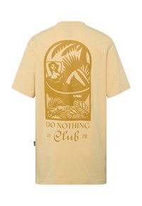 Camiseta de algodón amarilla con un gran gráfico marrón de una mujer descansando entre follaje tropical y el texto "Clube de No Hacer Nada".