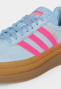 Φωτεινά μπλε αθλητικά παπούτσια Adidas Gazelle με έντονες ροζ ρίγες και λαστιχένια σόλα, με μπλε κορδόνια και "Gazelle" με χρυσά γράμματα.