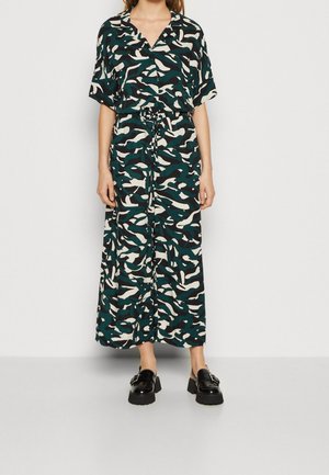 Kvinde iført en lang, løs jumpsuit med grønt, sort og hvidt abstrakt mønster og sorte chunky loafers.