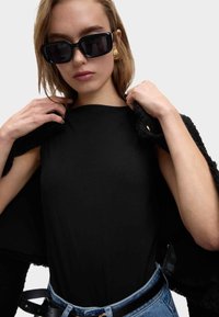 Vrouw met zwarte zonnebril, gouden oorbellen, zwarte top, blauwe jeans met riem, die een zwarte gestructureerde jas over haar schouders aanpast.