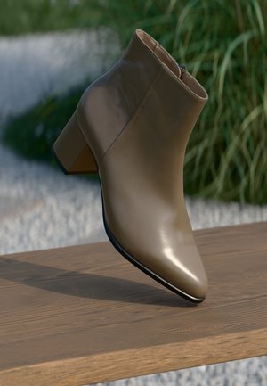 Beige leather ankle boot with a pointed toe and a chunky heel. Features a side zipper and smooth texture. Positioned above a wooden surface.