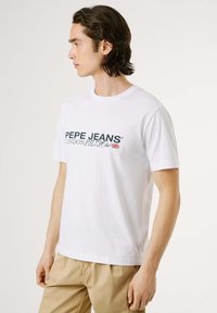 Balta īsām piedurknēm T-krekliņš ar "PEPE JEANS" logotipu tumši zilā krāsā, apaļas kakla izgriezums, brīvs piegriezums, izgatavots no kokvilnas, ar etiķeti apakšējā pusē.