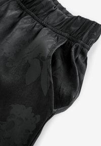 Mustat housut, joissa on kukkakuosi hieman tummemmassa sävyssä, joustava vyötärö ja sivutaskut. Materiaali näyttää sileältä ja kiiltävältä.