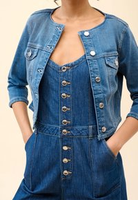 Nainen, jolla on päällään nappikiinnitteinen sininen farkkutakki, jossa on etutaskut, sekä lyhyt, kaulukseton farkkutakki hopeanvärisillä napeilla.