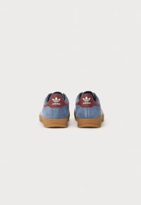 Zapatillas de ante azul con suela de goma marrón, acento en el talón de cuero burdeos y rayas laterales blancas. Presentan el clásico logo de Adidas en el talón.