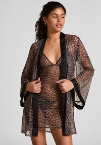 Vrouw in een doorschijnende jurk met luipaardprint en een bijpassende geur met zwarte afwerkingen, poseert met haar handen die de randen van de geur vasthouden, kijkend naar rechts.