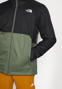 Chaqueta resistente al agua en negro y verde oliva, con cremallera frontal, bolsillos laterales y un logo en el pecho. Textura suave.