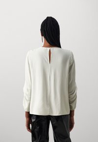 Blusa bianca a maniche lunghe con scollatura rotonda, dotata di un piccolo dettaglio a occhiello sul retro e realizzata in un tessuto morbido e liscio, abbinata a pantaloni neri lucidi.