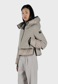 Giacca puffer corta beige con cappuccio, dotata di zip frontale, orlo regolabile e tasche laterali, abbinata a pantaloni grigi chiari.