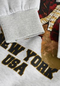 Jasnoszara bluza z ribbowanymi mankietami, z grafiką i czarnym oraz złotym napisem "NEW YORK USA" z przodu. Miękka w dotyku.