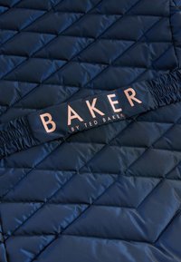 Gewatteerd marineblauw jack met diamantenpatroon, voorzien van een zwarte elastische band met 'BAKER' en 'by TED BAKER' in rosegouden letters gedrukt.