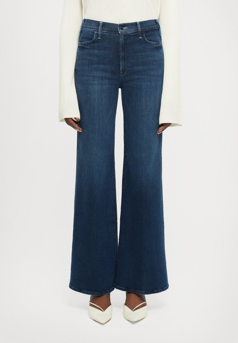 Jeans a vita alta, larghi e di colore blu scuro, indossati con tacchi bianchi a punta e un maglione a maniche lunghe color crema con maniche lunghe.