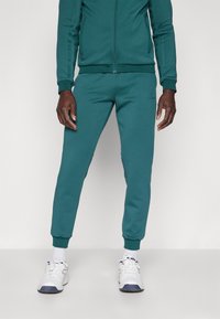 Teal treningsdrakt med glidelåsjakke og ribbestrikkede mansjetter samt matchende smale bukser. Materialet ser glatt ut med en myk tekstur.