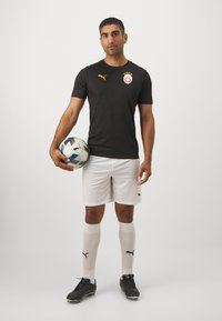 Maglia da performance nera con motivo testurizzato, dettagli arancioni, pantaloni corti bianchi, scarpe nere e calze bianche fino al ginocchio con logo Puma.