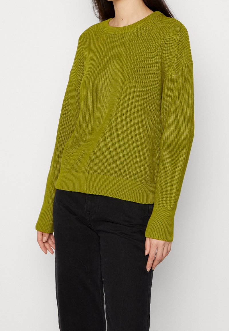 Personne portant un pull en laine vert mousse à manches longues associé à un pantalon noir sur un fond blanc uni.