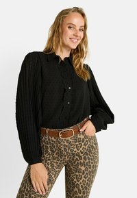 Blusa negra con lunares texturizados, borde de encaje detallado y mangas abullonadas. Combinada con pantalones ajustados de estampado de leopardo y un cinturón marrón.