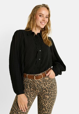 Blusa negra con lunares texturizados, borde de encaje detallado y mangas abullonadas. Combinada con pantalones ajustados de estampado de leopardo y un cinturón marrón.