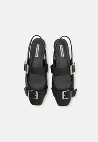 Zapatos de tacón negro tipo slingback con punta cuadrada, textura de cuero suave, hebillas metálicas prominentes y correas ajustables.