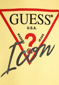 Camicia di cotone gialla con un logo triangolare rosso con scritto "GUESS" e "Icon" impreziosito da strass e testo nero al centro.