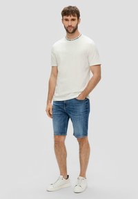 Biała koszulka z okrągłym dekoltem i czarnymi napisami przy kołnierzu, w połączeniu z niebieskimi szortami dżinsowymi i białymi trampkami.